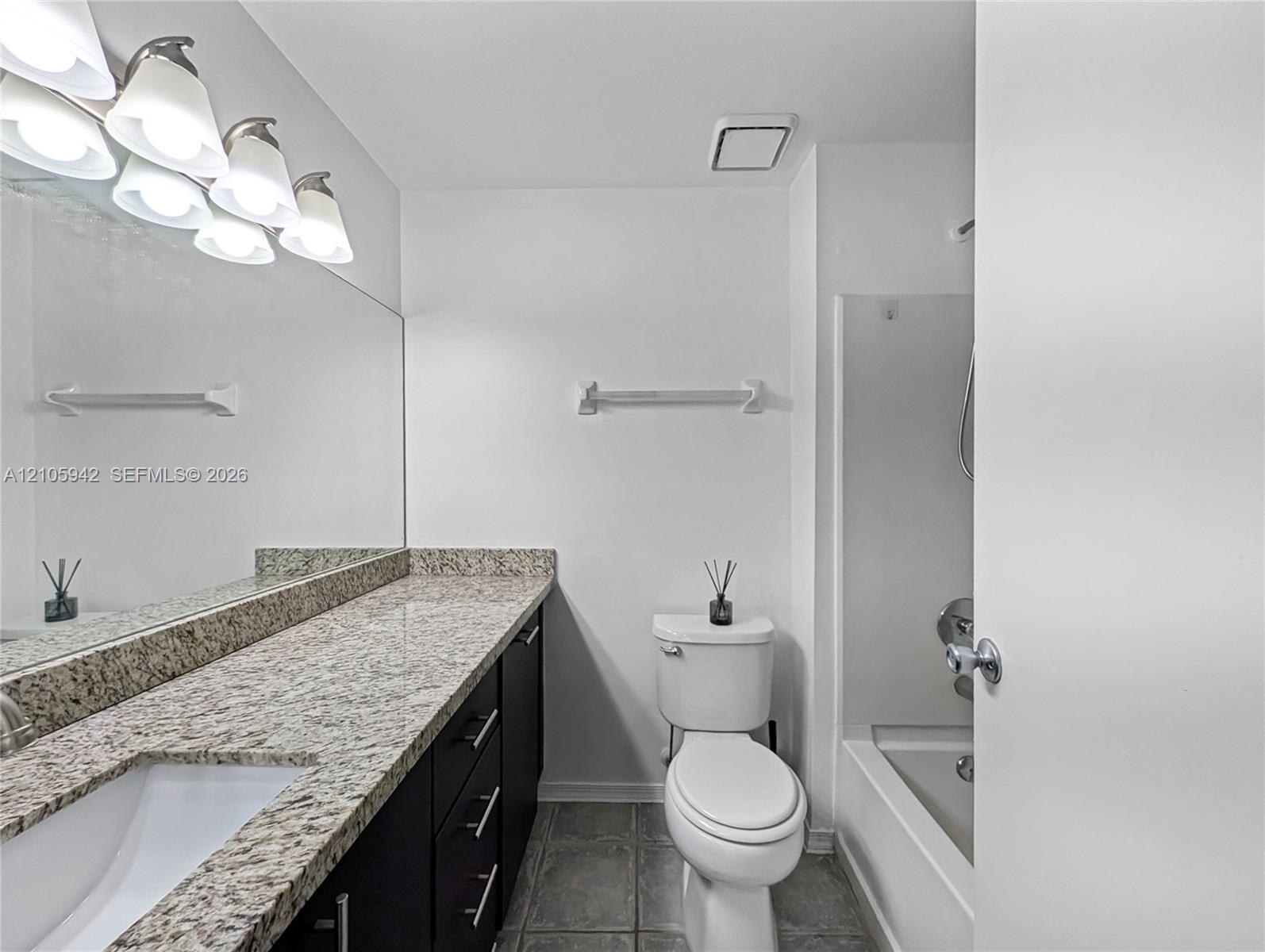 Full-width wall mirror, stone-composite countertop, dark-finish cabinetry, ceramic tile flooring, and a shower-tub combination