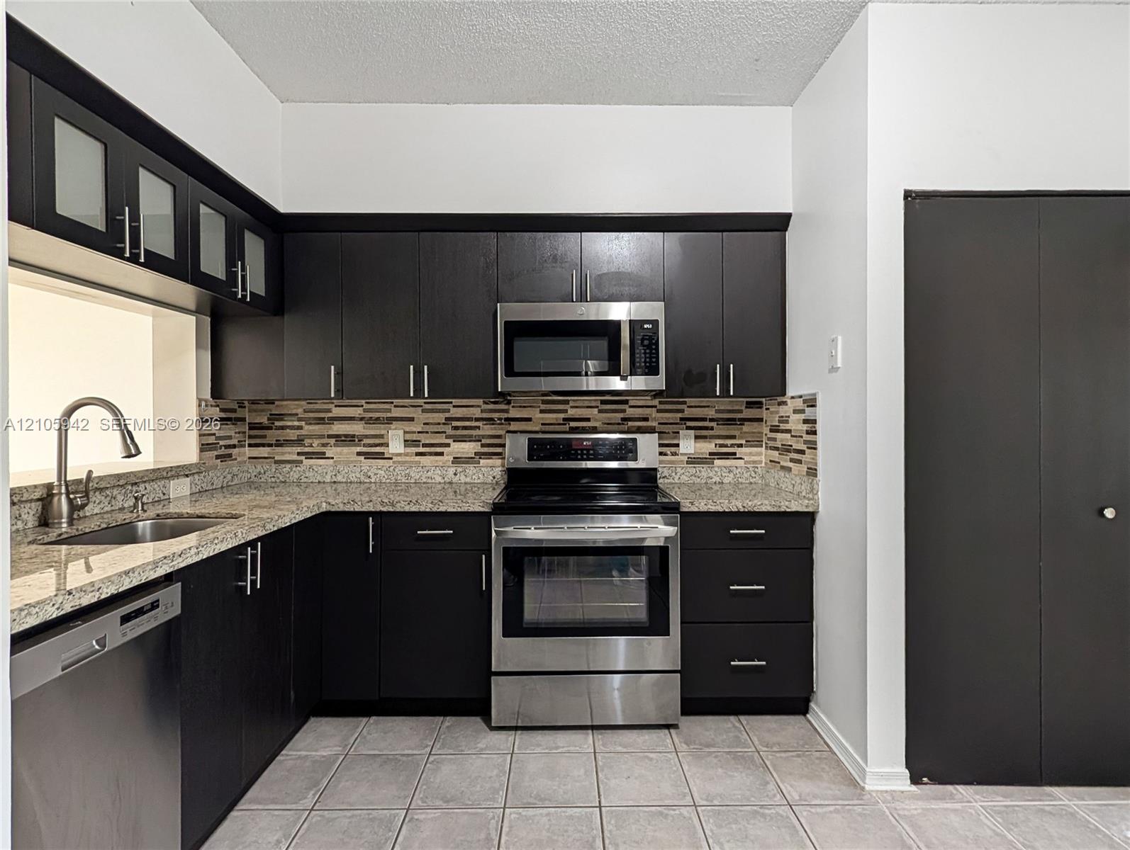 Dark cabinetry with glass-front upper accents, stone-composite countertops, stainless steel appliances, multi-tonal backsplash, and ceramic tile flooring