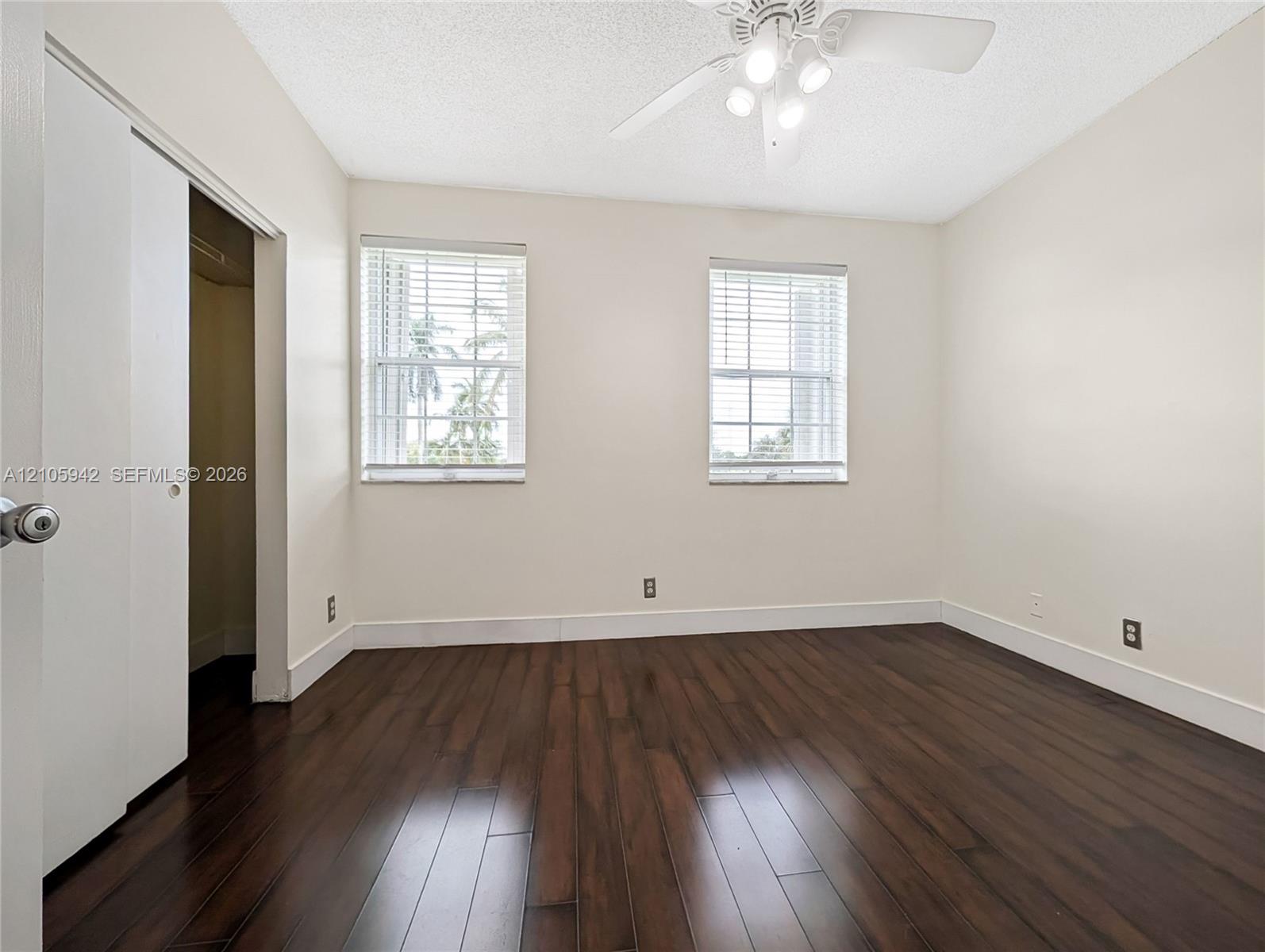 Room featuring dark-toned wood-finish flooring, two windows with horizontal blinds, and a white ceiling fan with integrated lighting