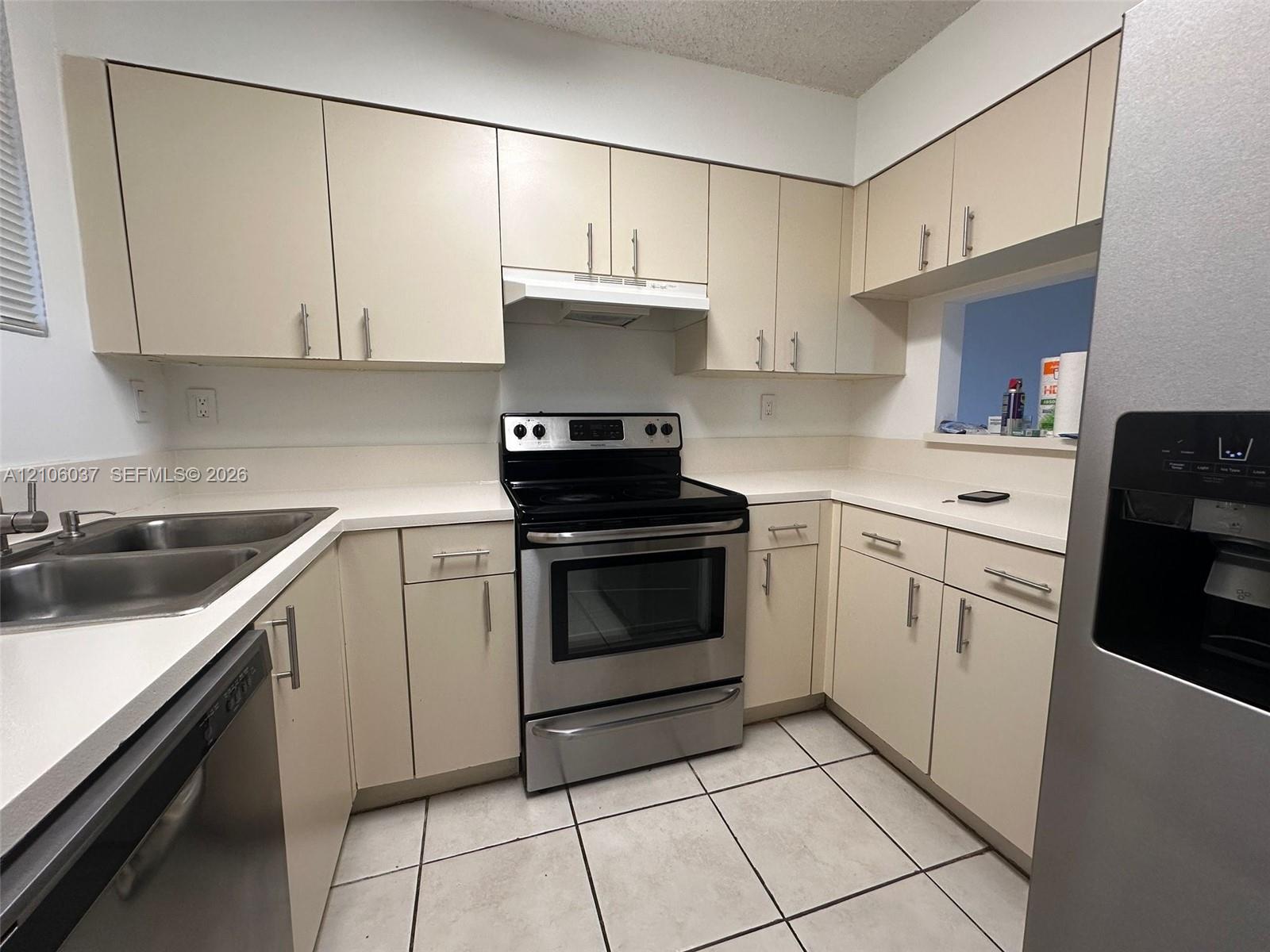 Kitchen featuring cream-toned cabinetry with metal hardware, solid-surface countertops, stainless steel appliances, a dual-basin sink, and tile flooring