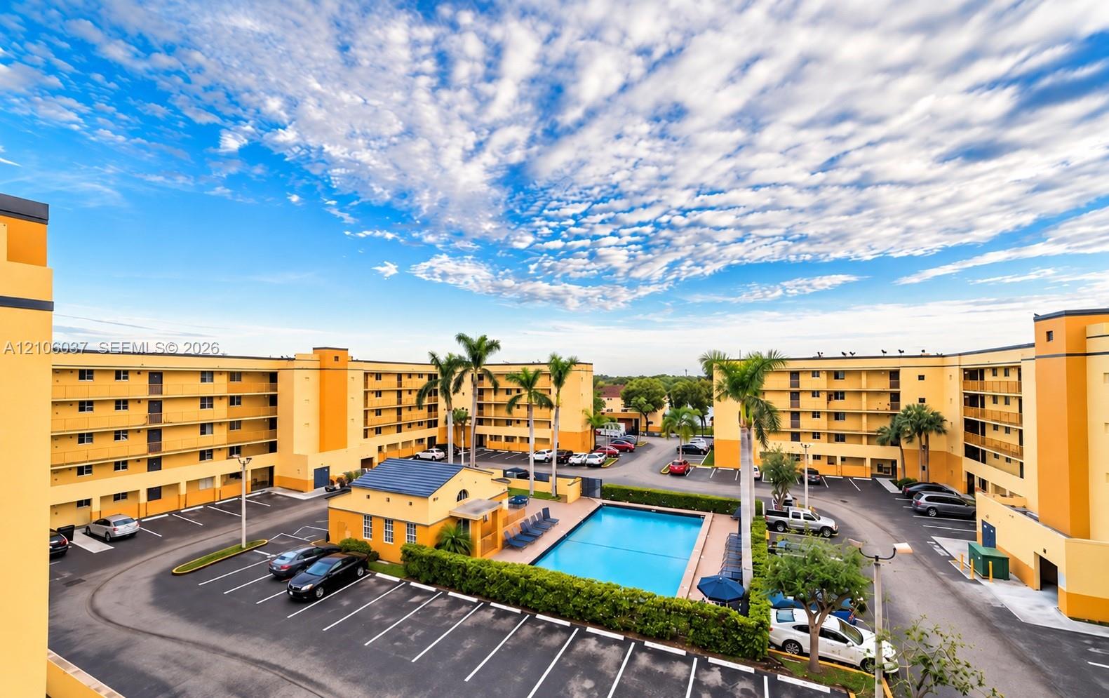 Multi-story residential complex featuring an outdoor swimming pool, paved surface parking, and mature palm trees