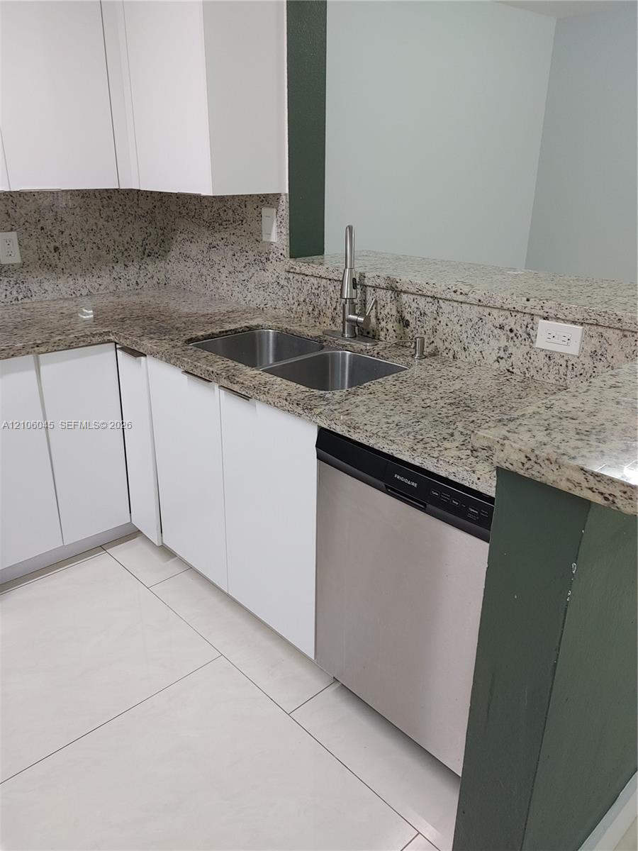 Kitchen featuring white cabinetry, stone countertops, double-basin stainless steel sink with high-arc faucet, stainless steel dishwasher, and large-format tile flooring