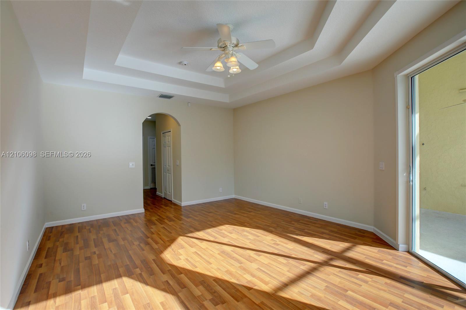 Room featuring a multi-tiered tray ceiling, wood-finish flooring, a central ceiling fan with multi-light fixture, an arched entryway, and a sliding glass door