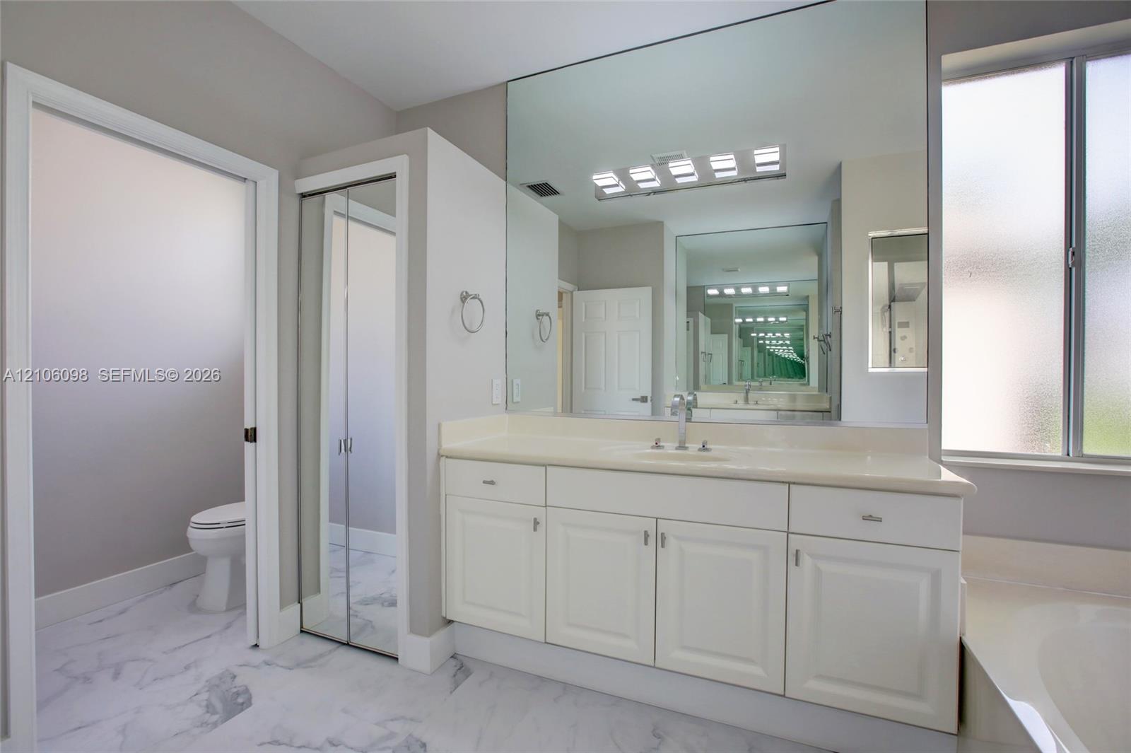 Bathroom featuring white cabinetry, solid-surface vanity countertop, full-width wall mirror, chrome-finish plumbing fixtures, and marble-patterned tile flooring