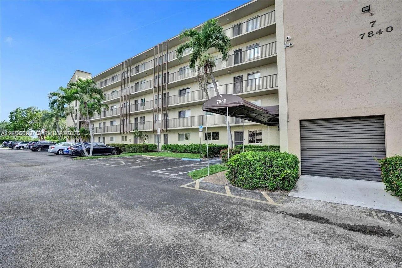 Multi-story stucco exterior building featuring balconies with metal railings, a covered entryway with a fabric awning, and surface-level parking