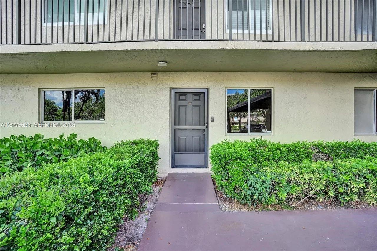 Stucco exterior featuring an entry door with a screen door, windows with aluminum frames, and a concrete walkway bordered by established shrubbery