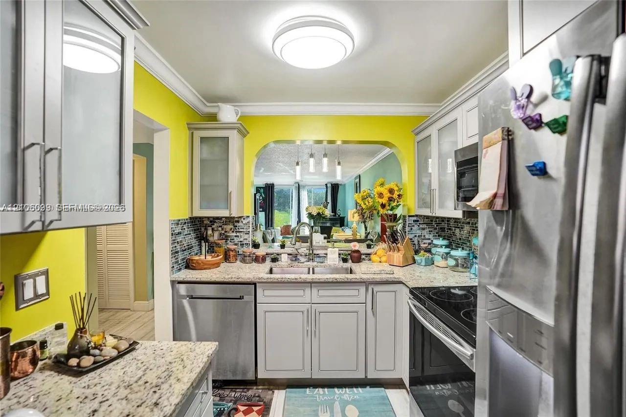 Kitchen featuring light-gray cabinetry, stone-finish countertops, stainless steel appliances, decorative tile backsplash, and a pass-through window with an arched header