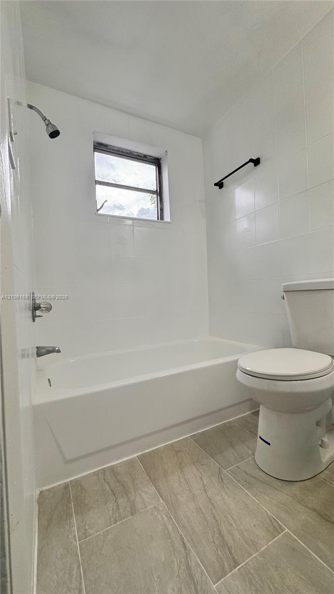 Bathroom featuring white wall tiling, a built-in tub with a shower head, ceramic tile flooring, and a matte black towel bar Bathroom featuring white wall tiling, a built-in tub with a shower head, ceramic tile flooring, and a matte black towel bar