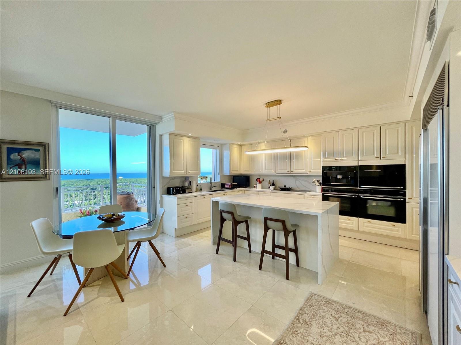 Kitchen featuring white cabinetry, an expansive center island with stone-finish countertops, built-in dual ovens, polished tile flooring, and sliding glass doors with ocean scenery