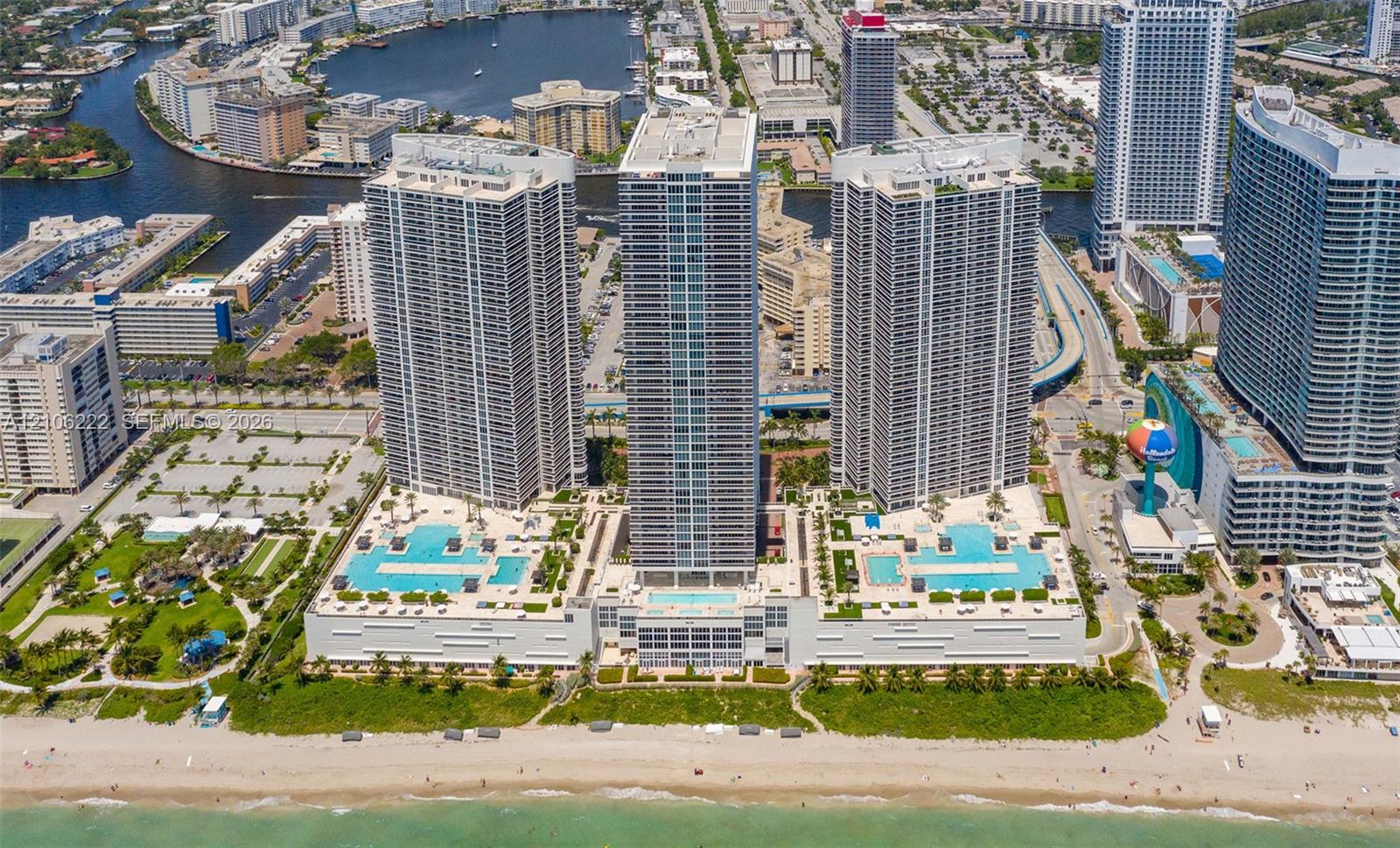 Aerial perspective of oceanfront high-rise residential complex featuring multiple swimming pools, spacious sun decks, extensive podium-level hardscaping, and direct beach access Aerial perspective of oceanfront high-rise residential complex featuring multiple swimming pools, spacious sun decks, extensive podium-level hardscaping, and direct beach access