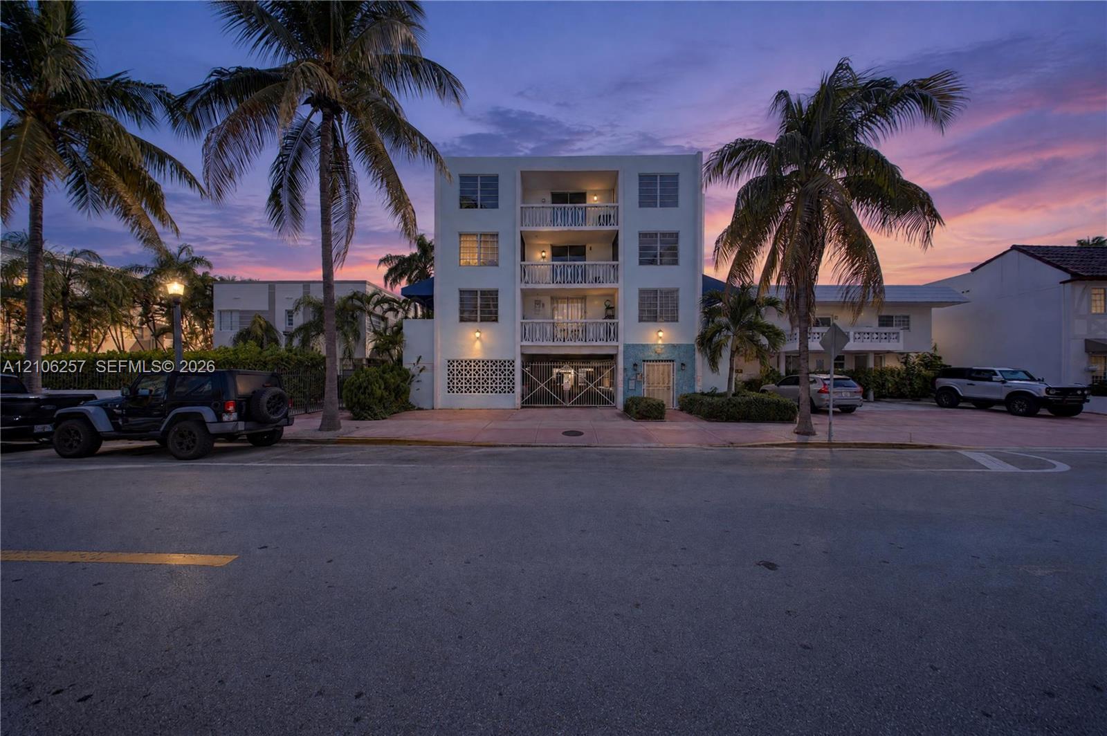 Multi-story building exterior featuring recessed balconies with decorative metal railings, geometric breezeblock accent wall, ground-level gated entry, and established palm trees, a couple of blocks away from the beach