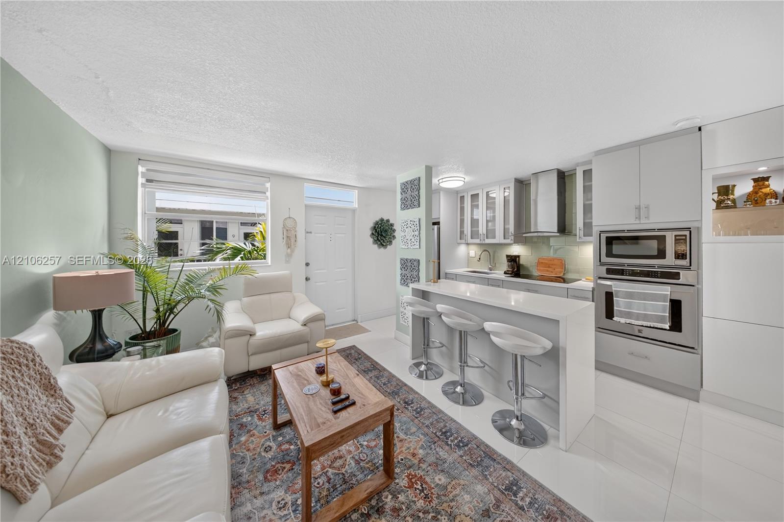Kitchen featuring light gray cabinetry, a breakfast bar with stone-style countertop, stainless steel built-in wall oven and microwave, stainless steel range hood, and tile flooring