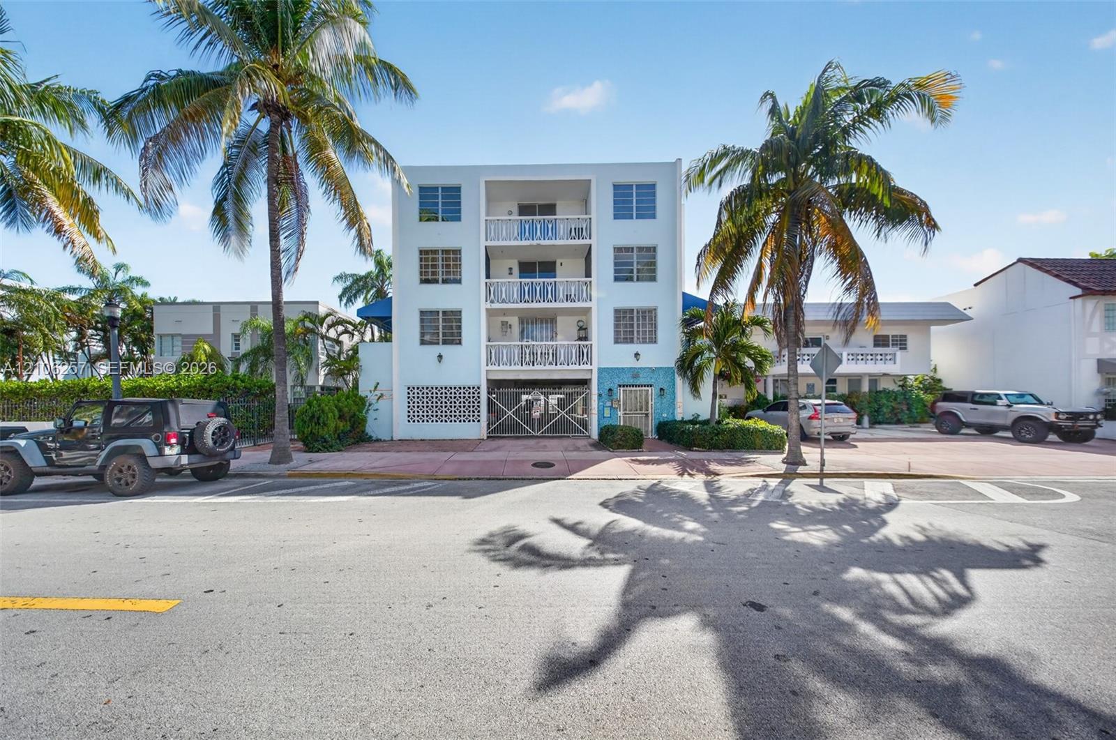 Multi-story residential building exterior featuring stacked balconies with patterned railings, decorative textured tile facade, louvered windows, and mature palm trees