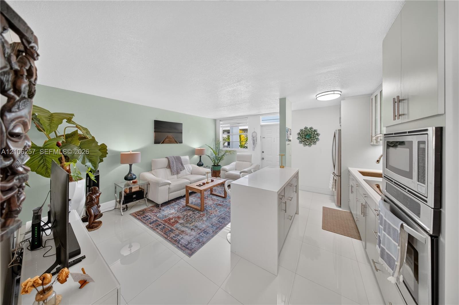 Open-concept living space featuring high-gloss tile flooring, white shaker cabinetry, a central island with a solid-surface countertop, and integrated stainless steel wall oven and microwave