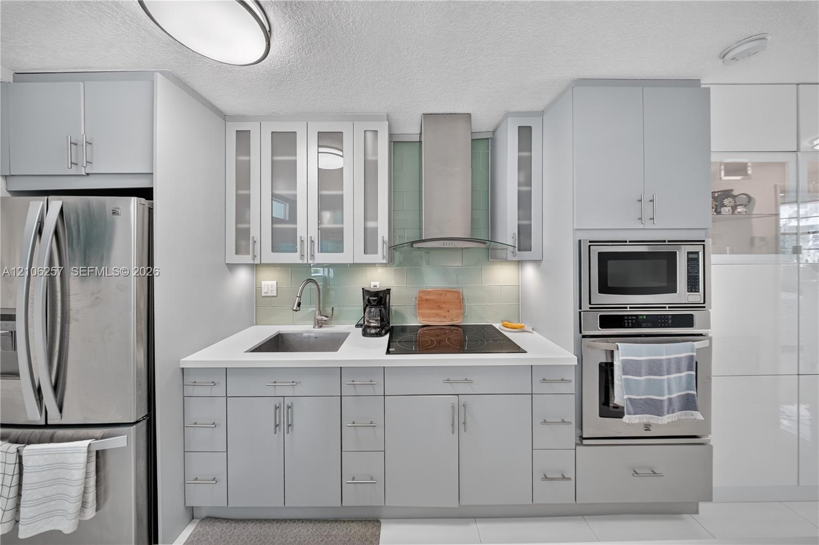 Kitchen featuring light gray cabinetry with glass-front uppers, solid-surface countertops, a glass tile backsplash, and stainless steel built-in appliances including a wall-mounted range hood
