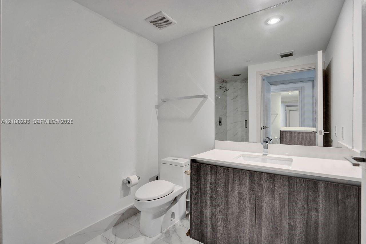 Modern vanity with dark wood-finish cabinetry, crisp white countertop, integrated rectangular sink, expansive wall-to-wall mirror, and stone-finish tile flooring