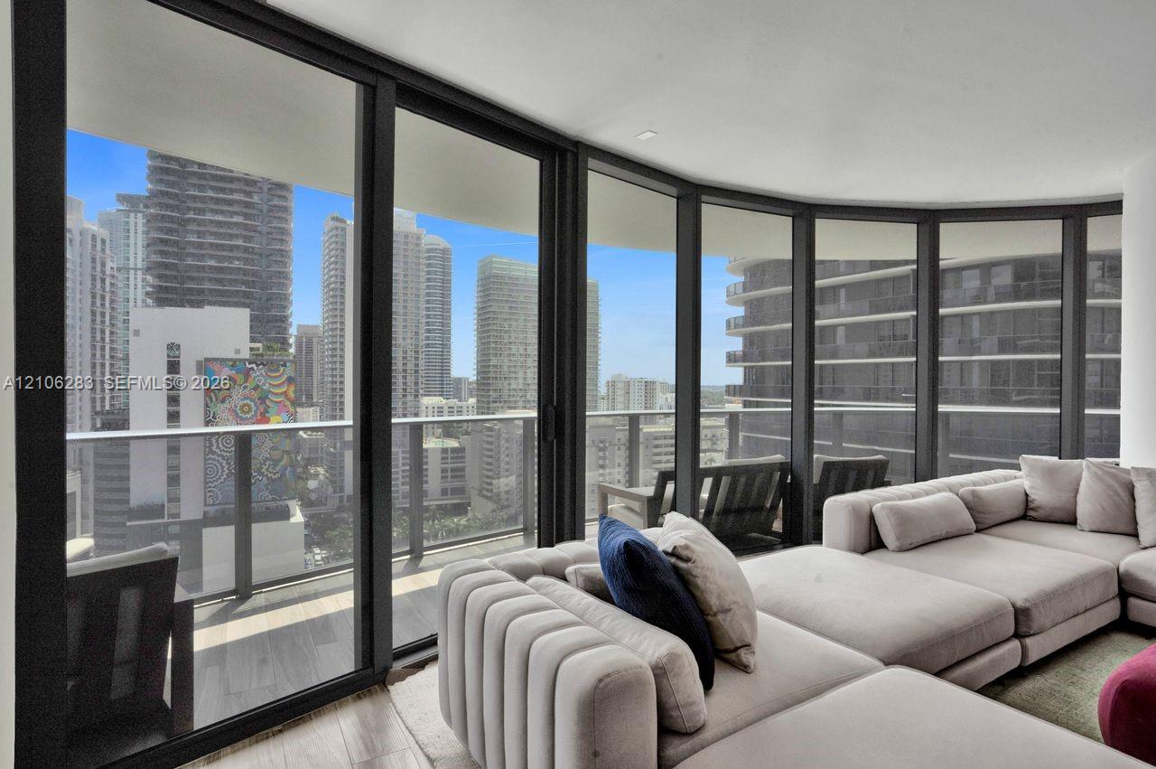 Floor-to-ceiling glass walls, wrap-around balcony with a metal railing, and neutral wood-finish flooring