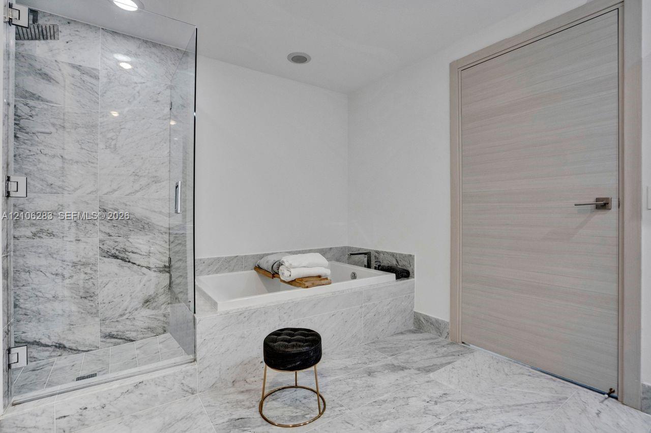 Frameless glass shower enclosure with stone tile surround, built-in soaking tub, stone tile flooring, and a light-toned wood-finish interior door
