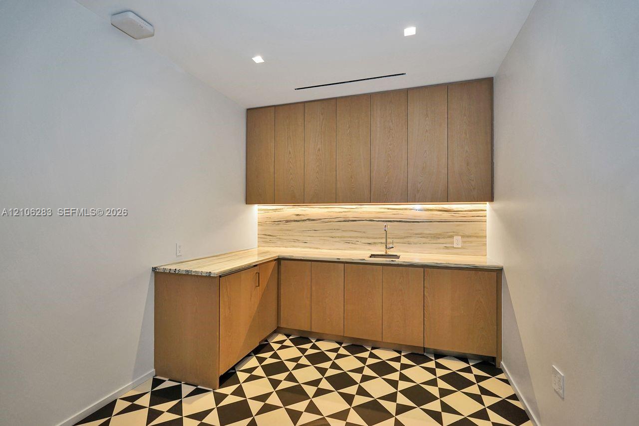 L-shaped cabinetry featuring flat-panel wood-finish doors, stone-material countertops and backsplash, integrated under-cabinet lighting, and a metal sink faucet with geometric patterned floor tile