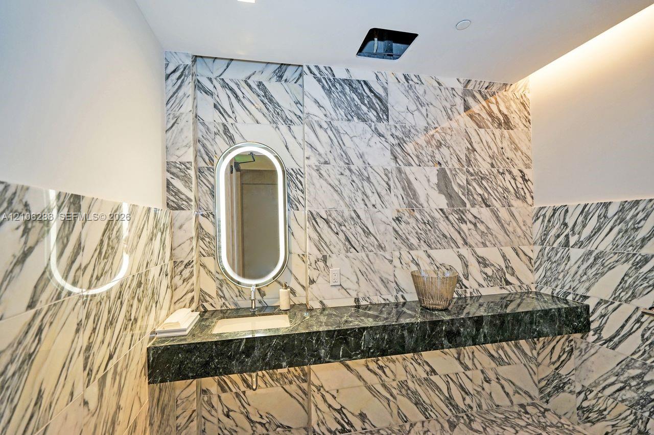 Floating vanity with a dark natural stone countertop, integrated sink, illuminated oval mirror, and floor-to-ceiling veined stone tile wall covering