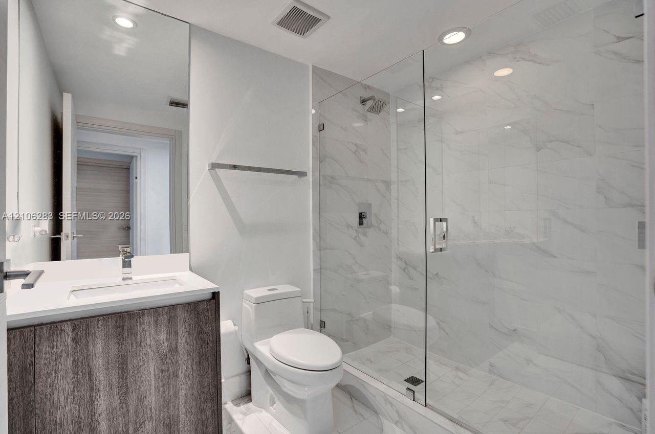 Bathroom featuring a vanity with wood-finish cabinetry, a white countertop, a walk-in shower with a glass enclosure, floor-to-ceiling stone-style tile, and recessed lighting