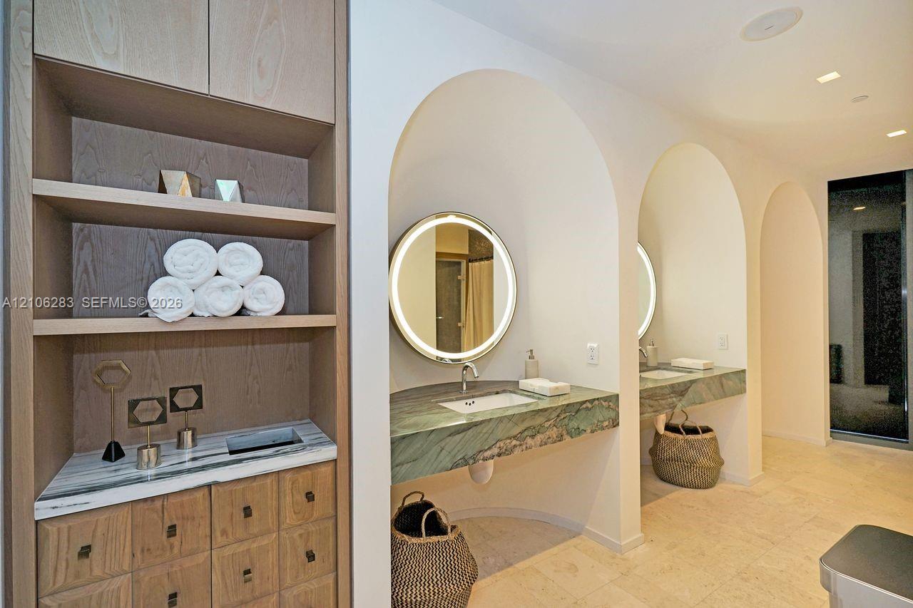 Custom wood-finish built-in cabinetry, stone-topped floating vanities recessed within arched alcoves, circular backlit mirrors, and natural stone tile flooring