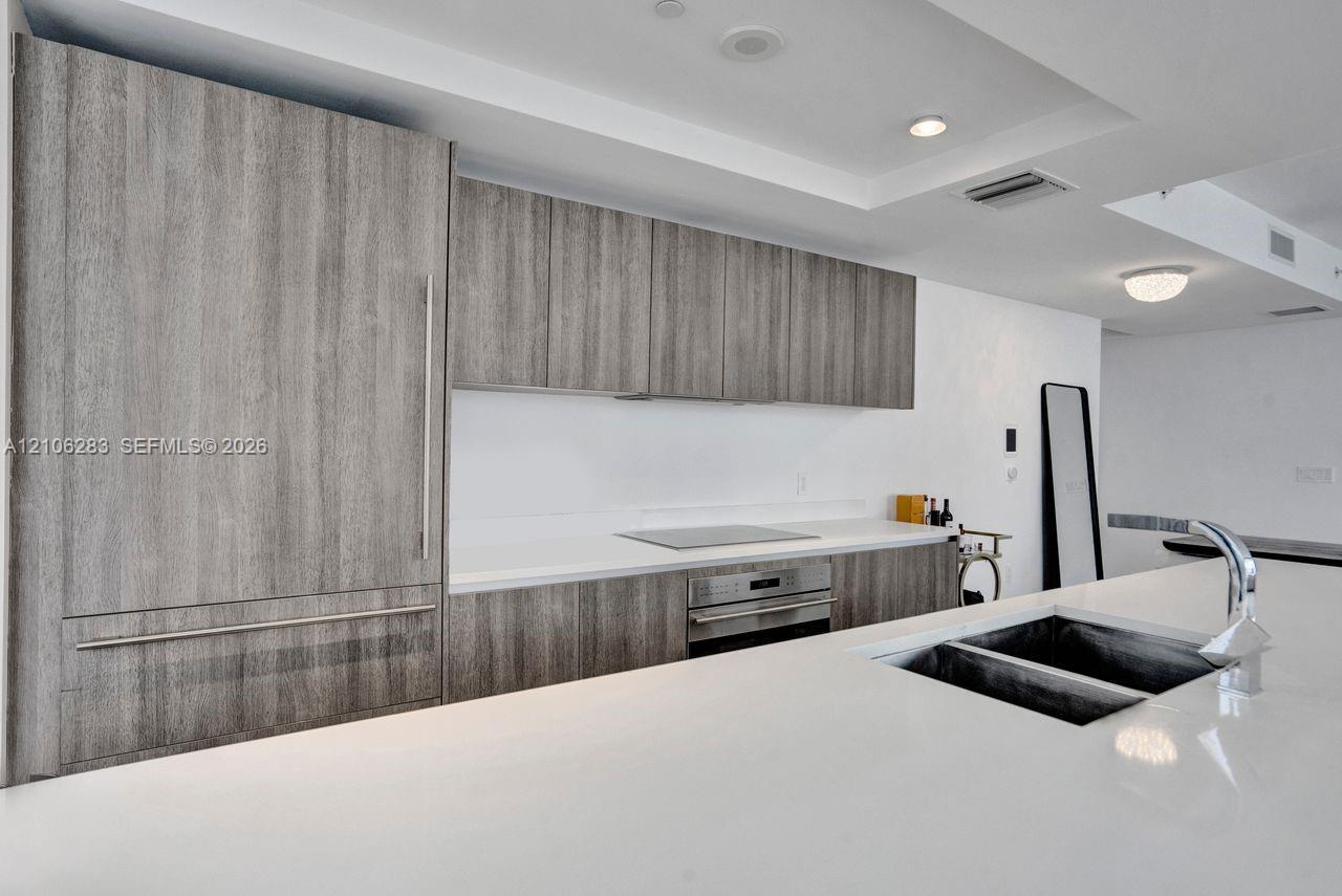 Kitchen featuring wood-finish cabinetry, integrated panel-ready refrigerator, solid-surface countertops, stainless steel wall oven and cooktop, and an undermount double-basin sink with a chrome faucet