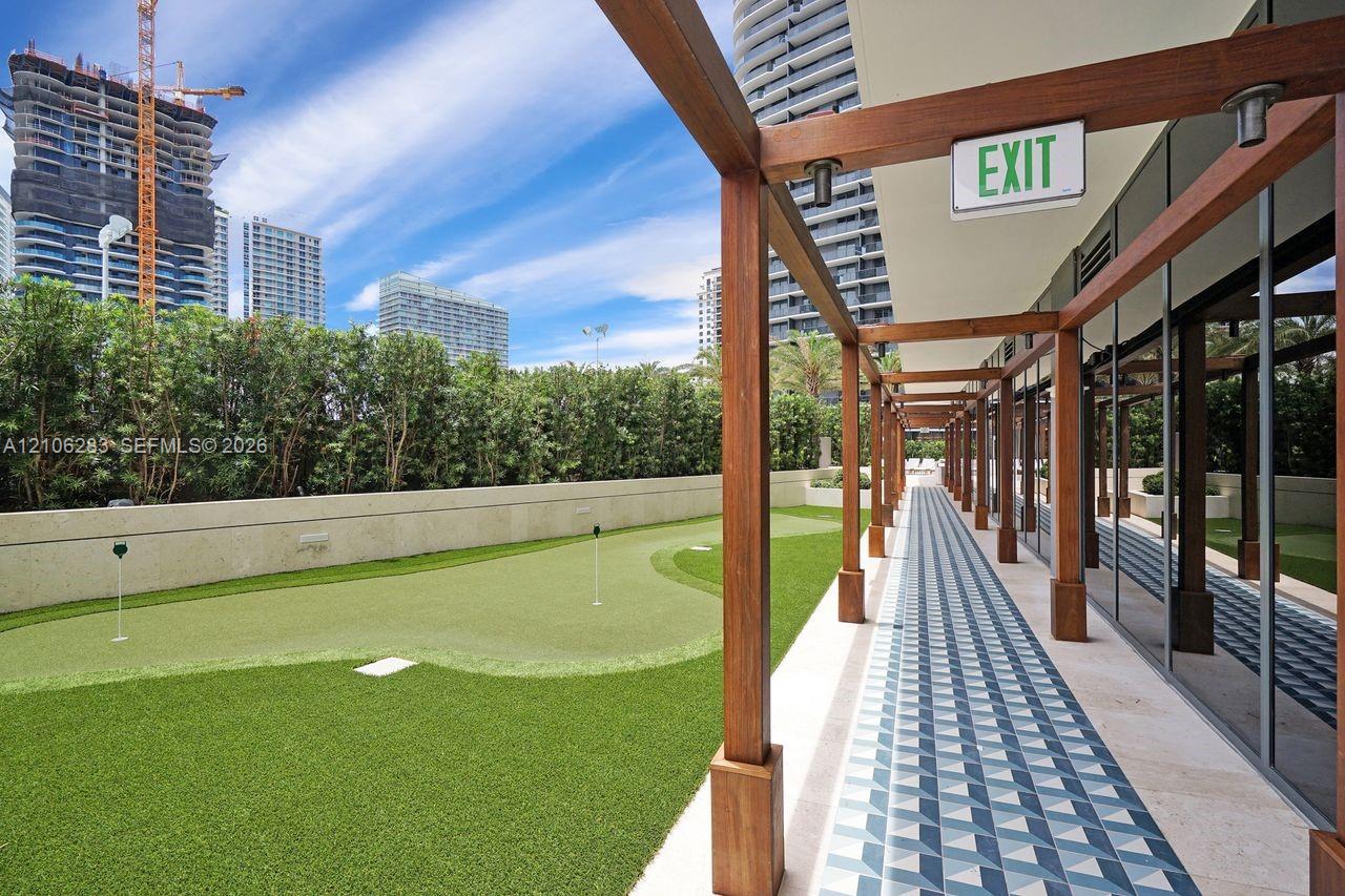 Outdoor putting green featuring artificial turf, bordered by a wood-framed pergola and patterned tile walkway with glass wall finishes