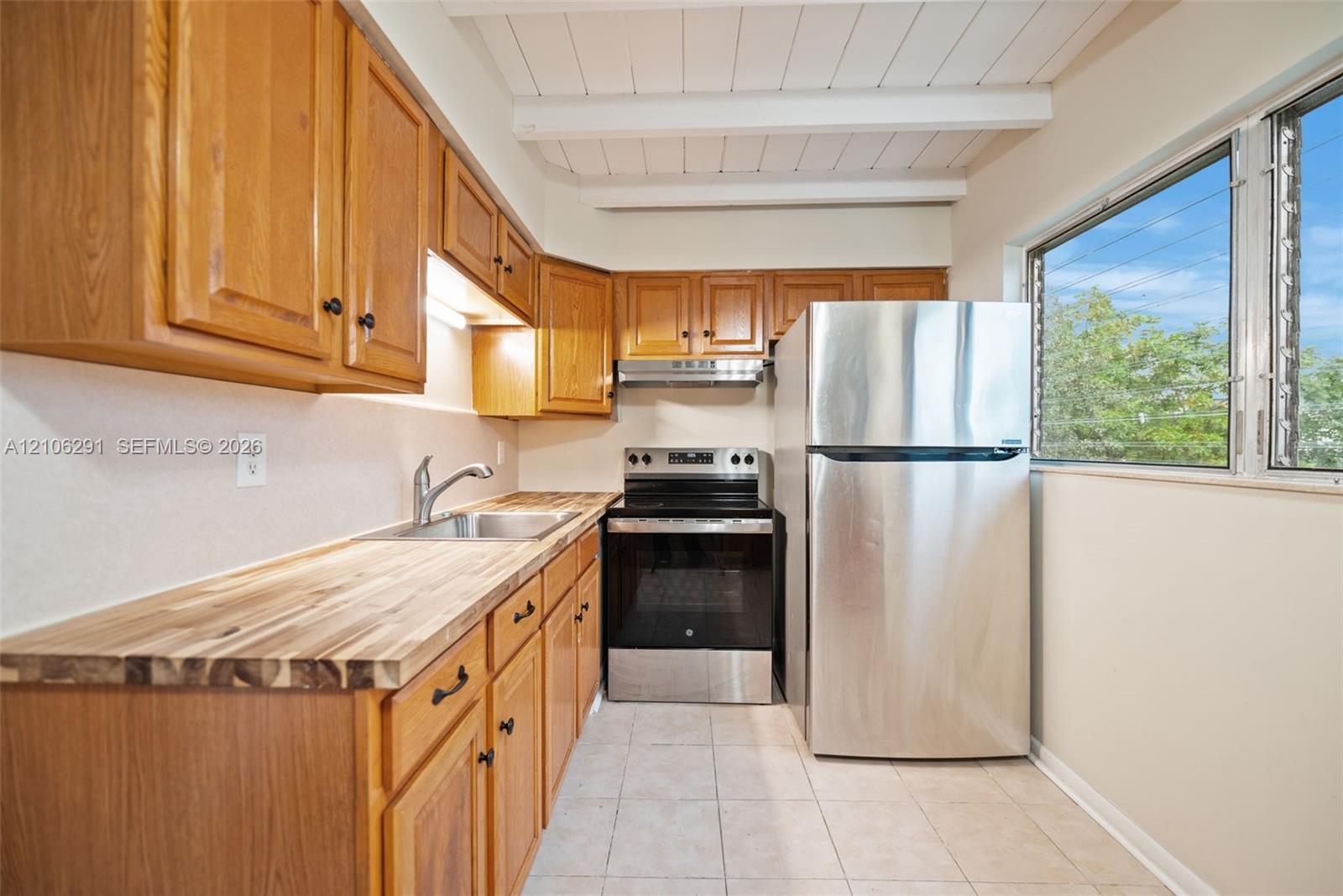 Kitchen featuring stained wood cabinetry, butcher-block countertops, stainless steel appliances, tile flooring, and a ceiling with exposed white beams