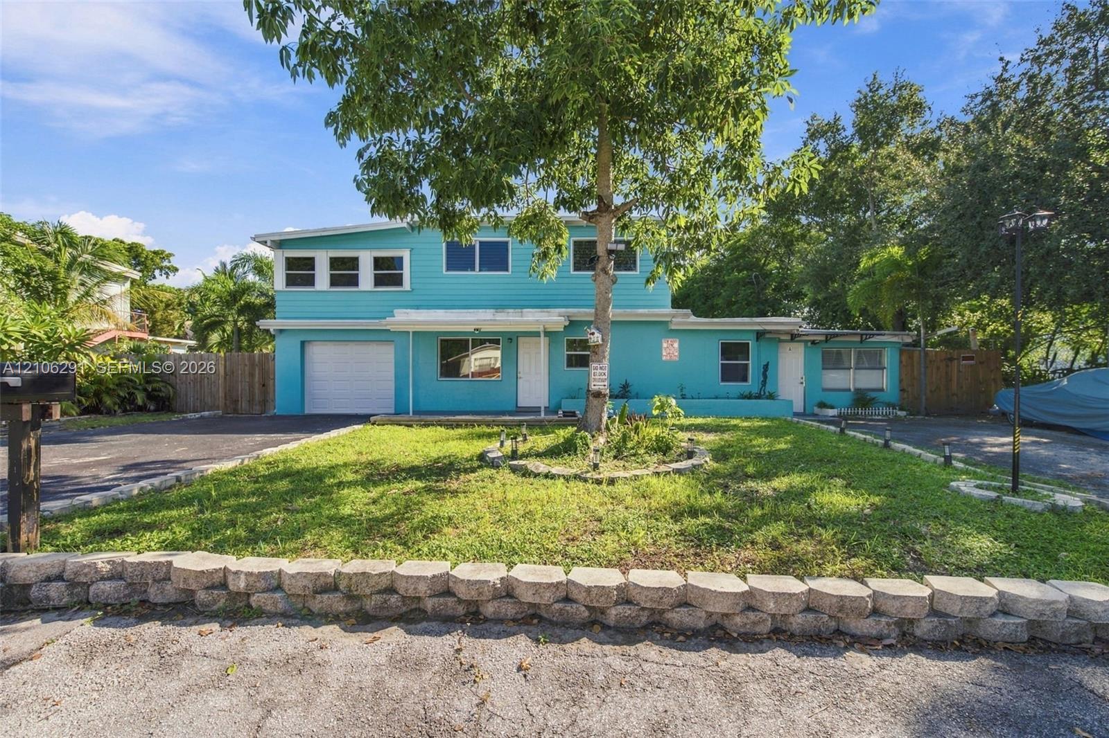 Two-story exterior with horizontal siding, attached garage, stone retaining wall, paved driveway, and mature landscaping