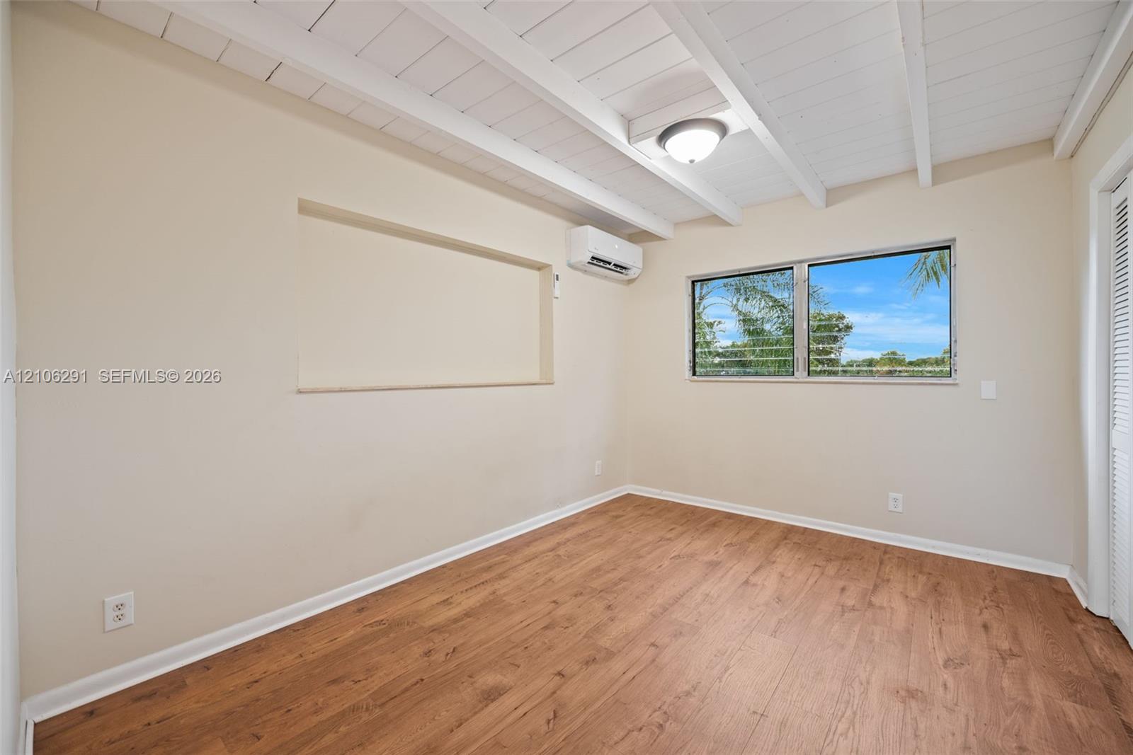Room featuring white exposed ceiling beams, wood-finish flooring, a wall-mounted ductless air conditioning unit, a recessed wall niche, and a window with horizontal panes