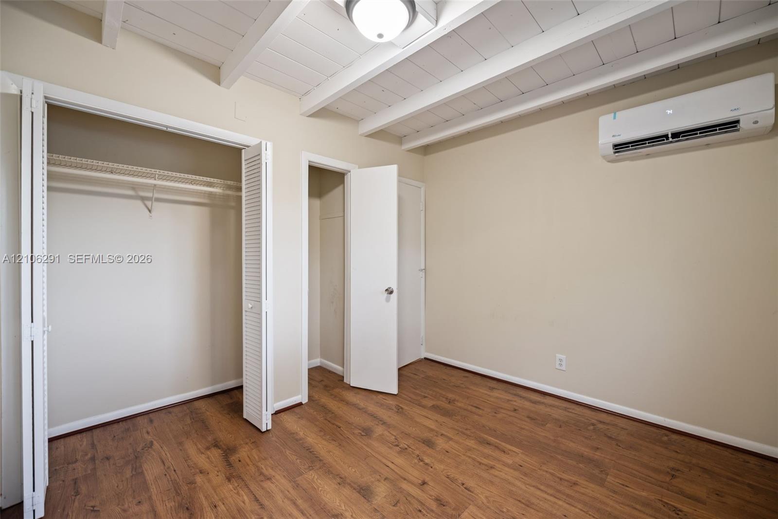 Room featuring wood-finish flooring, exposed painted ceiling beams, a wall-mounted ductless air conditioning unit, and dual closet storage options