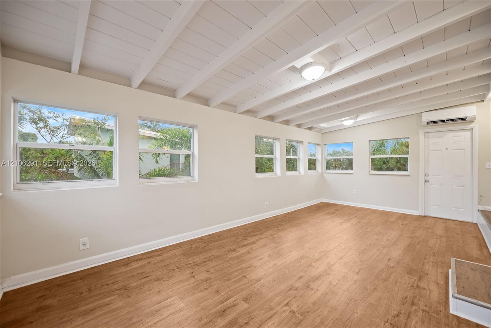 Interior room featuring wood-finish flooring, exposed white-painted beam ceiling, multiple windows, and wall-mounted climate control unit