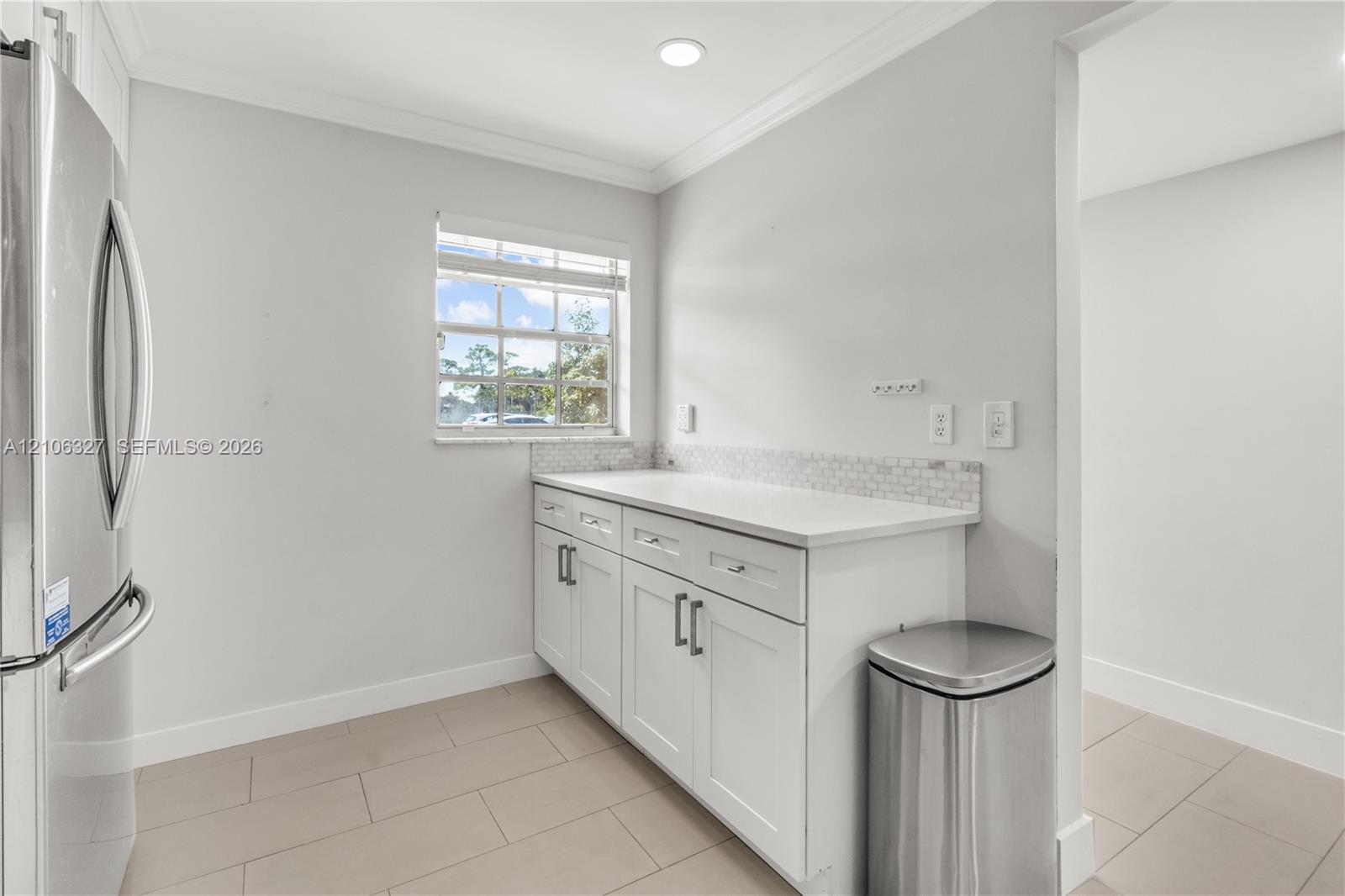 Shaker-style white cabinetry featuring chrome hardware, solid-surface countertop, mosaic tile backsplash, stainless steel refrigerator, and ceramic tile flooring