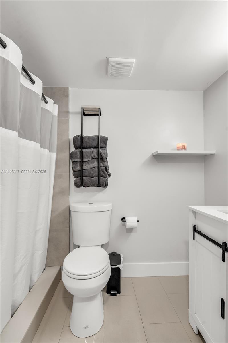 Bathroom featuring large-format neutral tile flooring, white vanity with matte black hardware, integrated tub surround with neutral accent tiling, and a wall-mounted floating shelf