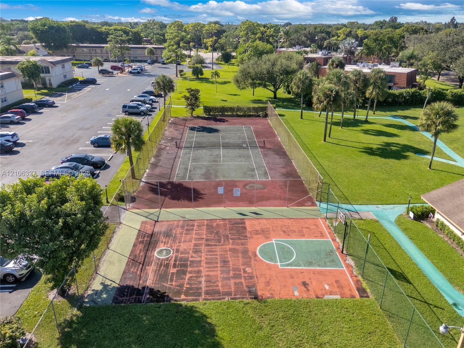 Outdoor recreation area featuring a fenced tennis court and a multi-purpose basketball court surrounded by manicured grounds