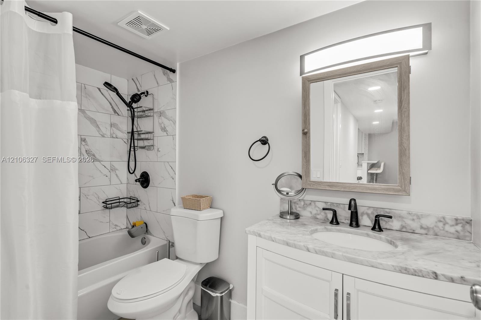 Bathroom featuring a stone-finish countertop with an integrated oval basin, white shaker-style cabinetry, a light-toned wood-frame medicine cabinet, and a bathtub enclosure with marble-patterned wall tile and a matte black shower fixture