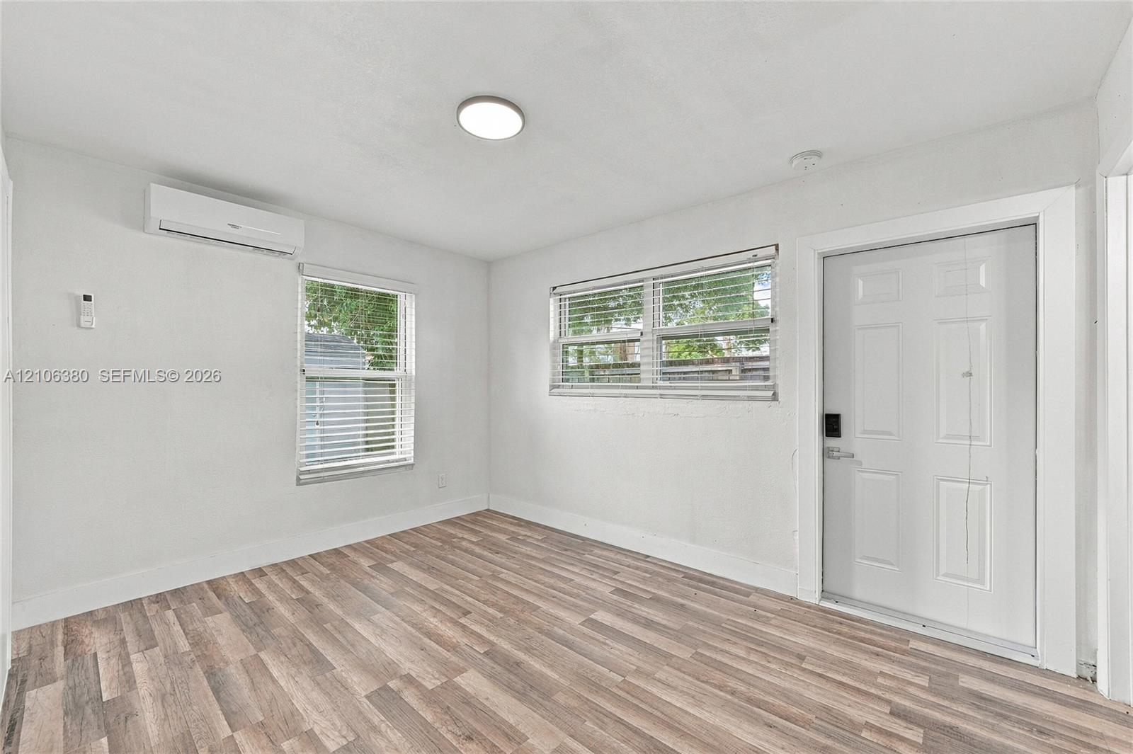 Wood-finish flooring, white walls, dual windows with horizontal blinds, wall-mounted ductless air conditioning unit, and white six-panel exterior door