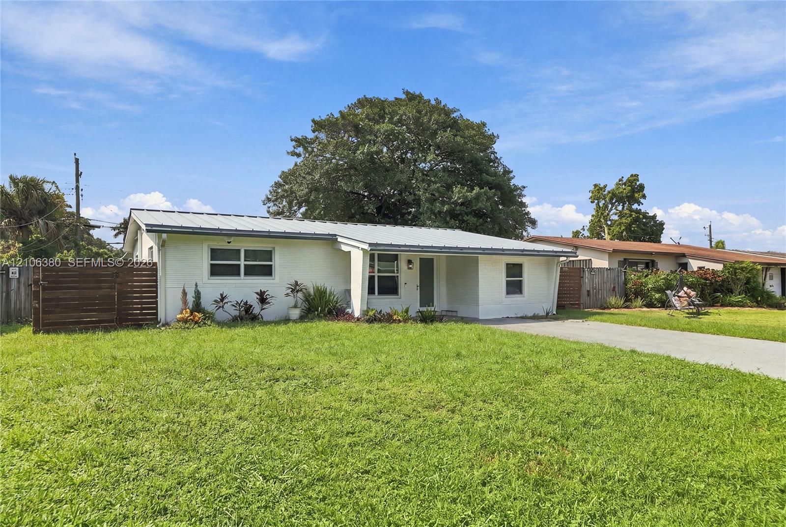 Single-story residence featuring a metal roof, white exterior cladding, concrete driveway, horizontal wood-slat privacy fencing, and expansive lawn