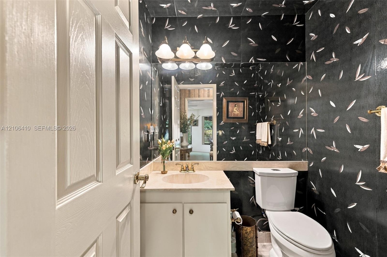 Powder room featuring patterned wall covering, stone-finish countertop with an integrated basin, vanity cabinet with dual-door storage, full-width vanity mirror, and three-light wall-mounted fixture