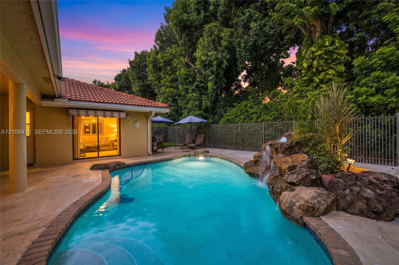 In-ground swimming pool featuring a naturalistic rock waterfall, stone-tile decking, and surrounding metal fencing against a backdrop of mature trees