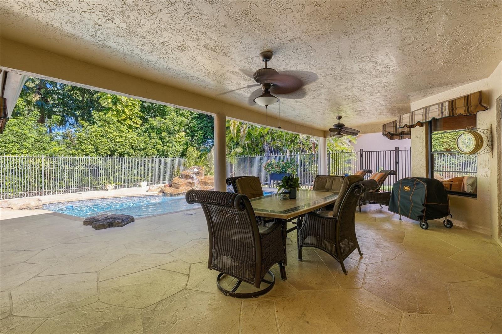 Covered outdoor patio featuring stone-patterned concrete flooring, two ceiling fans, a swimming pool with a rock water feature, and a metal perimeter fence