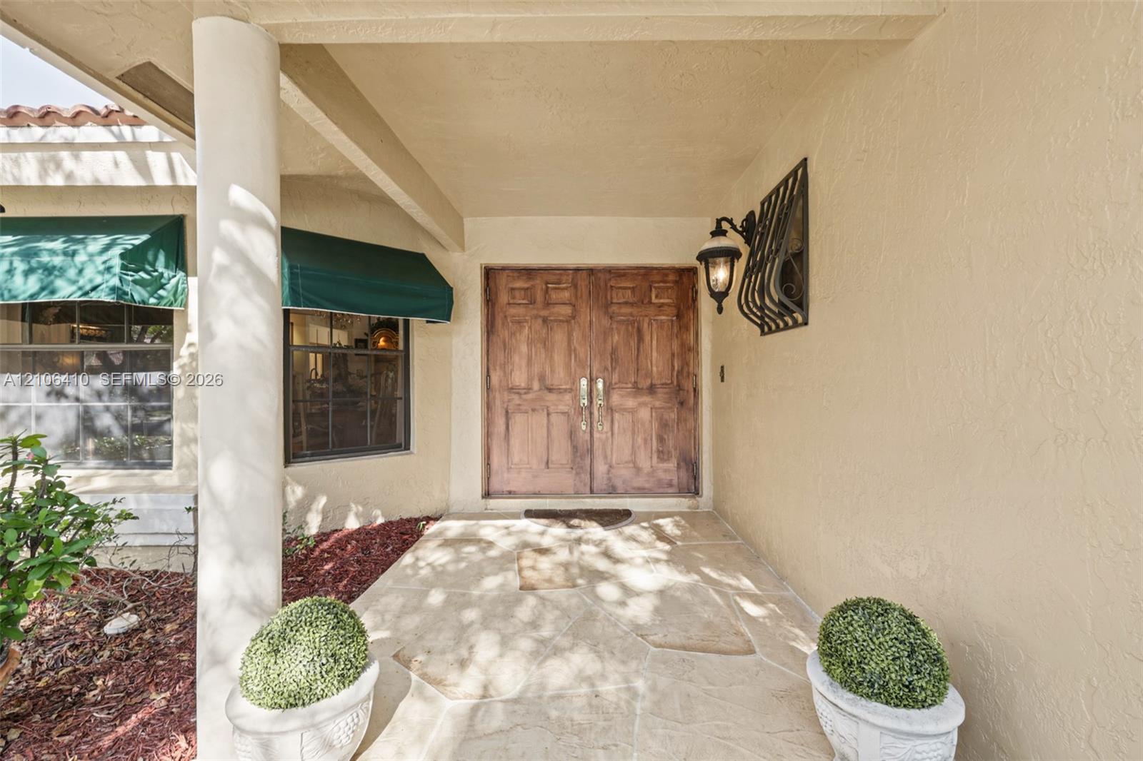 Covered entryway featuring flagstone paving, double stained-wood doors, decorative wall-mounted lighting, and fabric window awnings