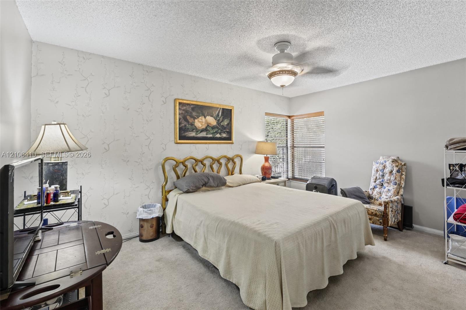Bedroom featuring neutral wall-to-wall carpeting, an accent wall with patterned wall covering, a multi-blade ceiling fan with integrated lighting, and horizontal window blinds