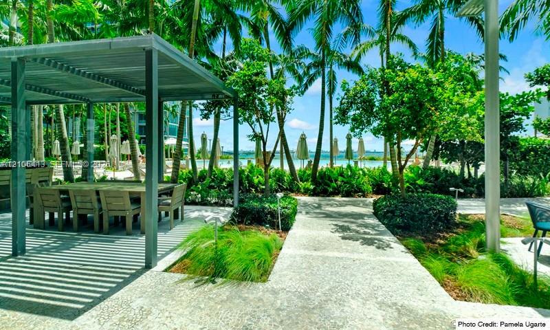 Outdoor patio area featuring a metal-frame pergola, stone-paved pathways, professional landscaping with mature palms, and built-in outdoor kitchen cabinetry