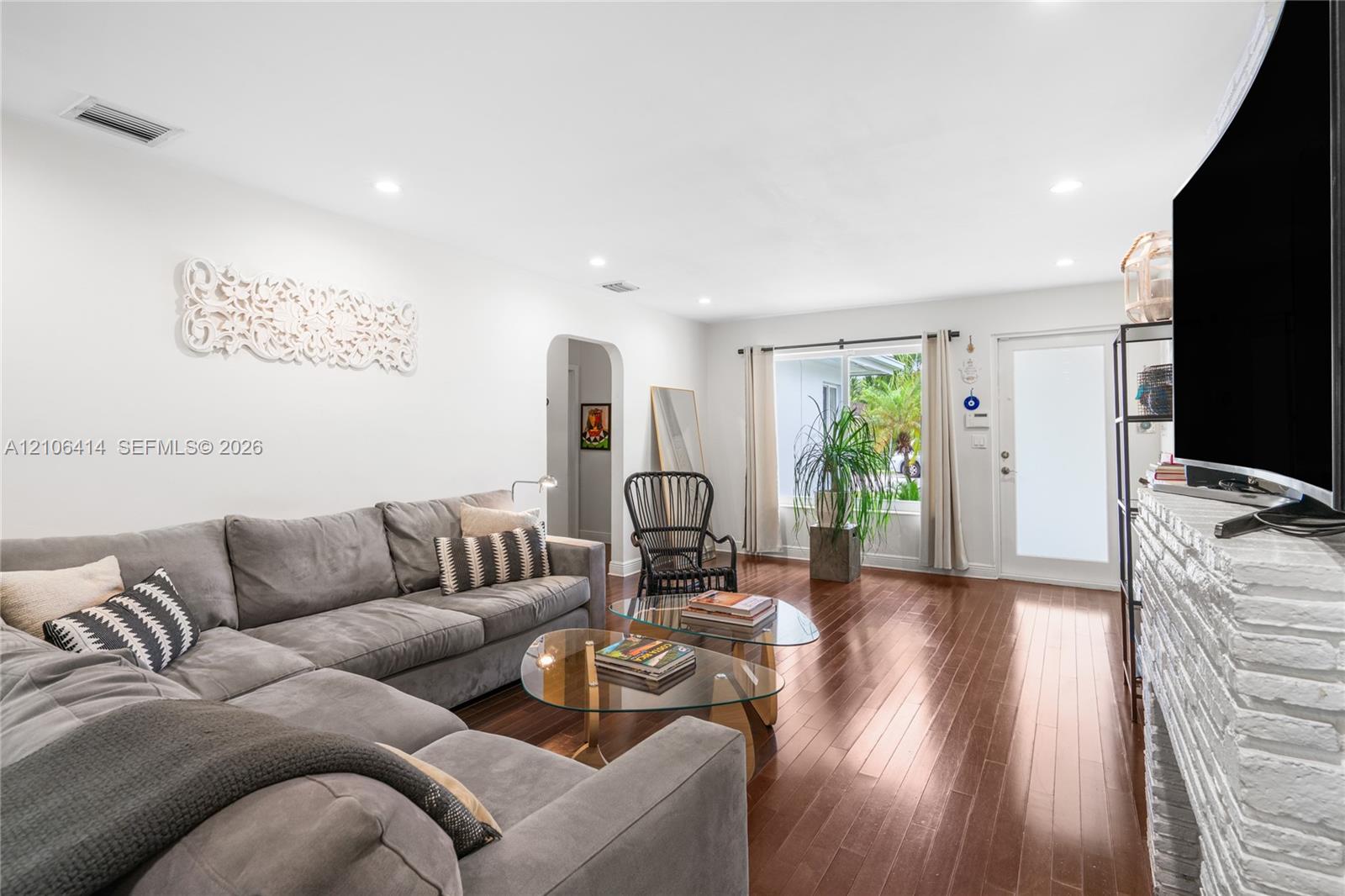Living area featuring wood-finish flooring, white-painted brick fireplace, recessed lighting, arched entryway, and a glass-panel entry door