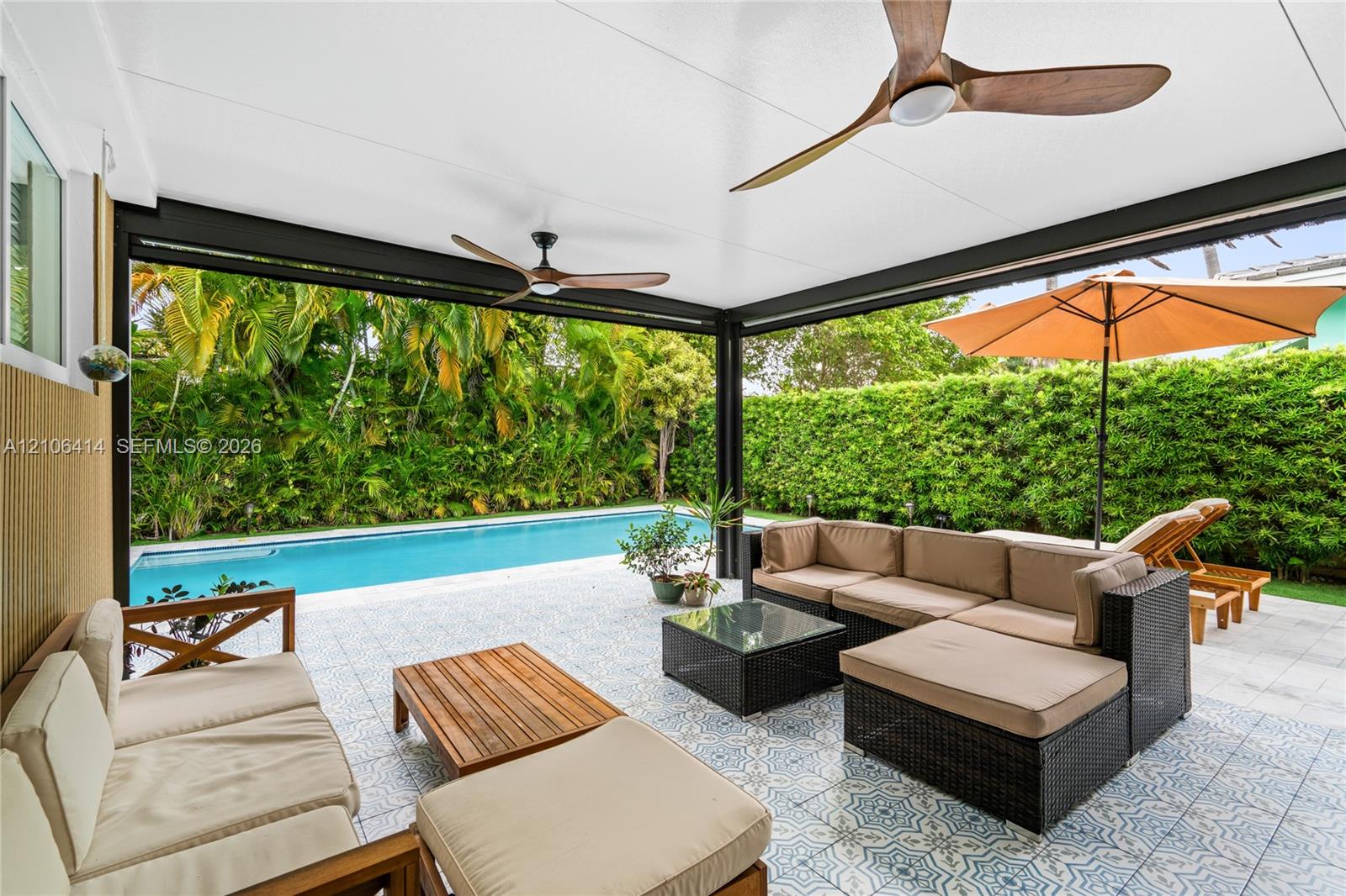 Covered outdoor patio featuring patterned tile flooring, dual wooden-blade ceiling fans, and a rectangular swimming pool surrounded by a tall hedge
