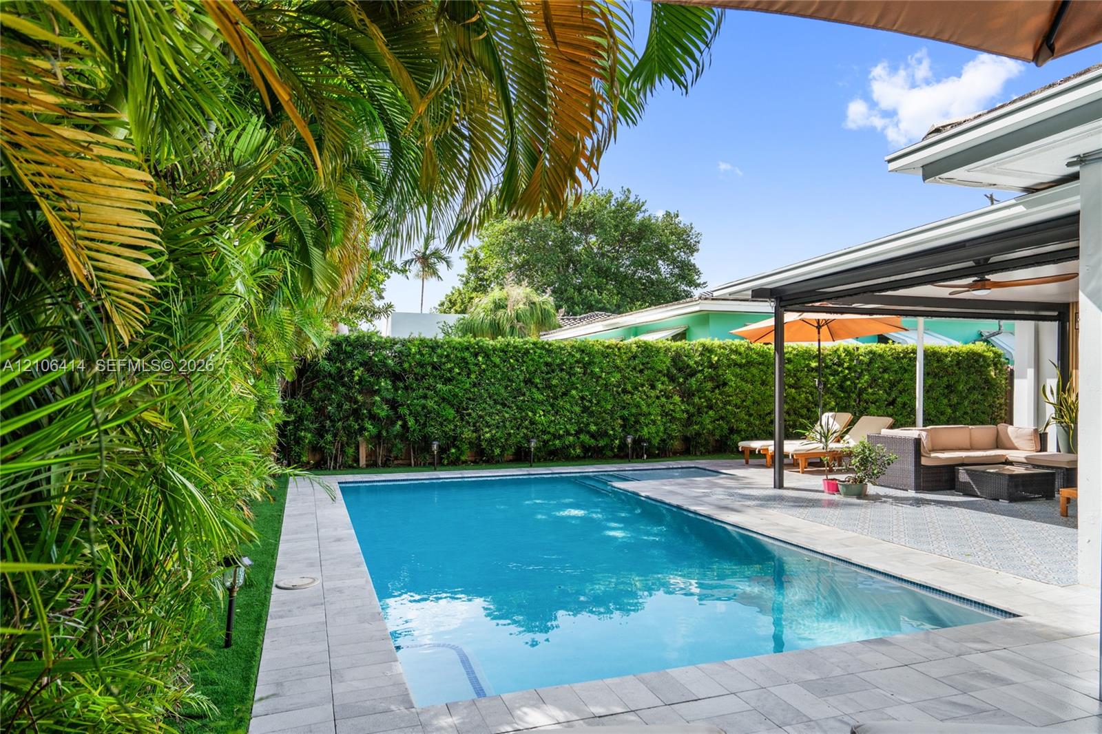 In-ground swimming pool featuring a stone-paver deck, covered lanai, and surrounding privacy hedges
