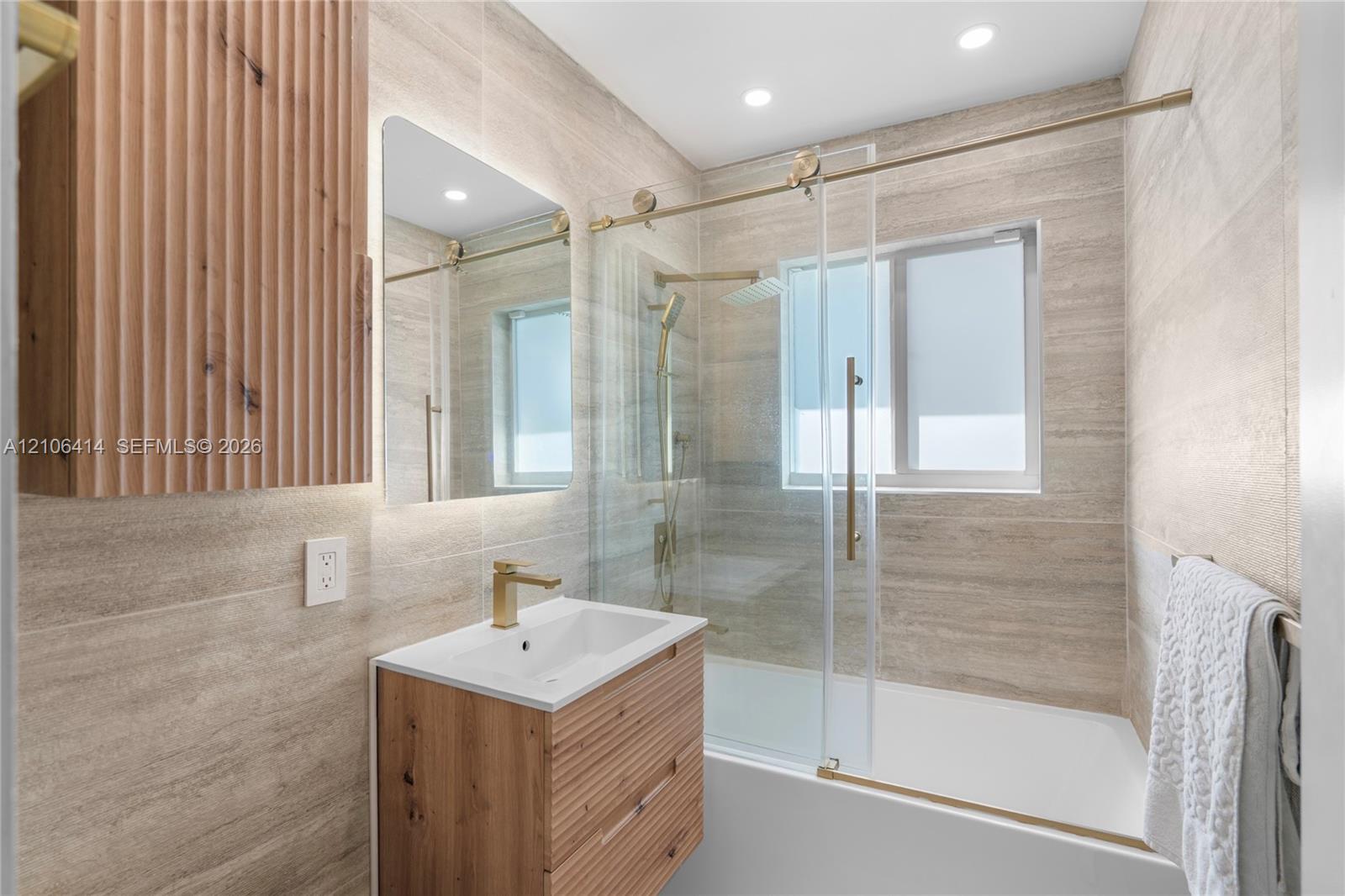 Bathroom featuring floor-to-ceiling stone-look tiling, a slatted wood-finish vanity with an integrated basin, gold-tone hardware, and a glass-enclosed bathtub
