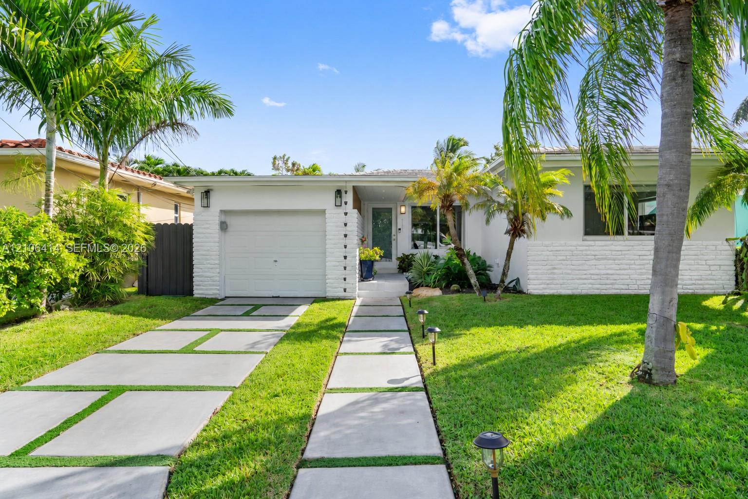 Single-story residence featuring white painted brick masonry, a concrete paver driveway, a manicured lawn, and mature palm trees